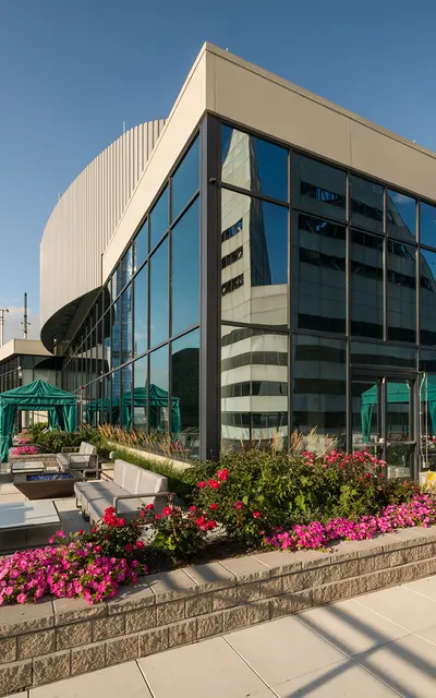 A panoramic view of a modern rooftop terrace with lounge furniture and flowering plants in the foreground, featuring large glass windows of a building and a tall skyscraper in the background under a clear blue sky.