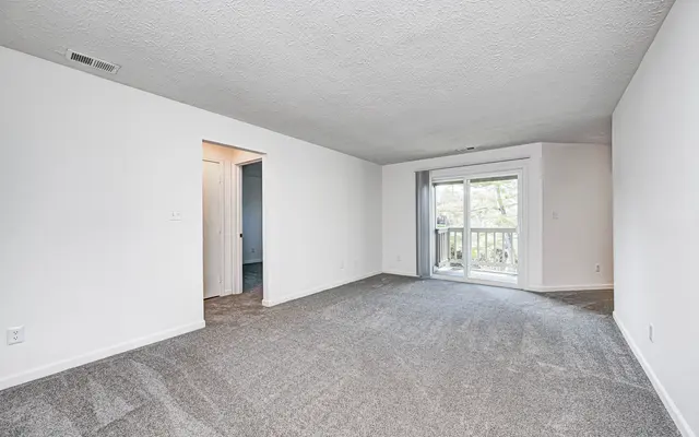 A spacious empty living room with carpeted flooring, a sliding glass door leading to a balcony, and a doorway to another room.