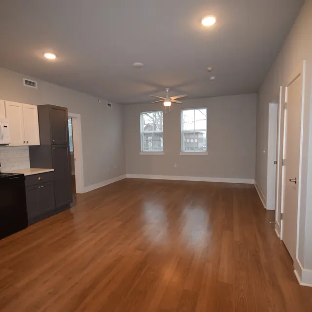 An open living space featuring a modern kitchen with white cabinets and black appliances, a wooden floor, and large windows allowing natural light.