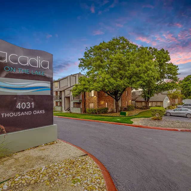 Entrance sign for Acadia on the Lake with apartment complex and trees in the background under a colorful sky.