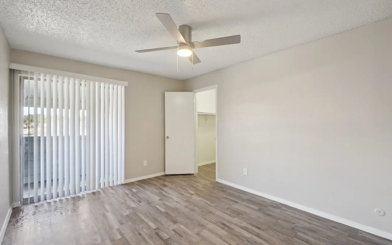 An empty room with light-colored walls and wooden flooring, featuring a ceiling fan and a set of vertical blinds on a glass door leading to a balcony.