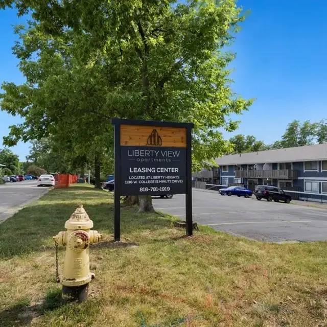 A sign for Liberty View Leasing Center by a fire hydrant on a lawn with trees and vehicles in the background.