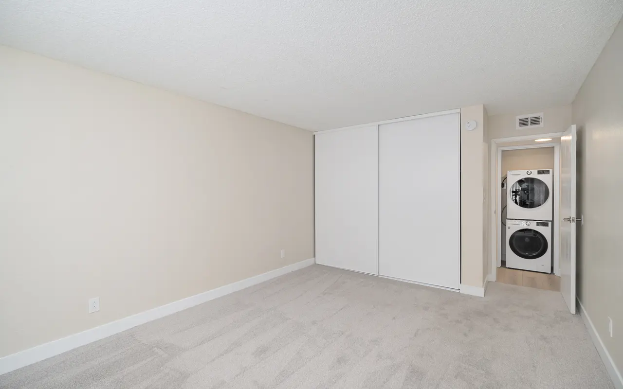 A spacious, empty bedroom featuring beige walls, soft carpet flooring, and a closet with sliding doors. In the corner, there is a stackable washer and dryer, with a doorway leading to another room.
