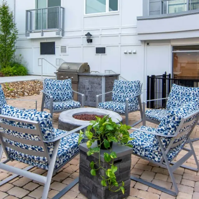 A cozy outdoor seating area featuring six chairs with blue patterned cushions arranged around a fire pit, with a grill nearby and a garden space visible in the background.