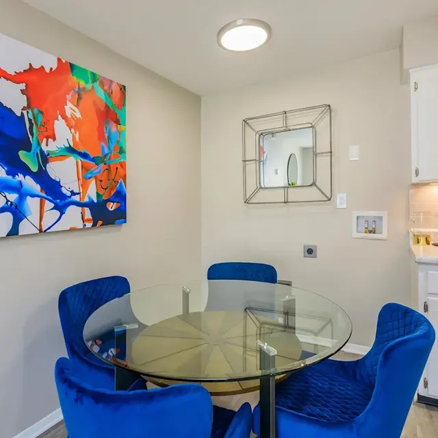 Bright dining area with a glass table, blue chairs, colorful abstract artwork on the wall, and a modern mirror.