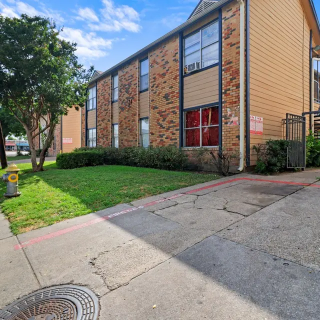 Exterior view of a two-story apartment complex with brick and wooden siding, surrounded by grass and trees, on a sunny day.