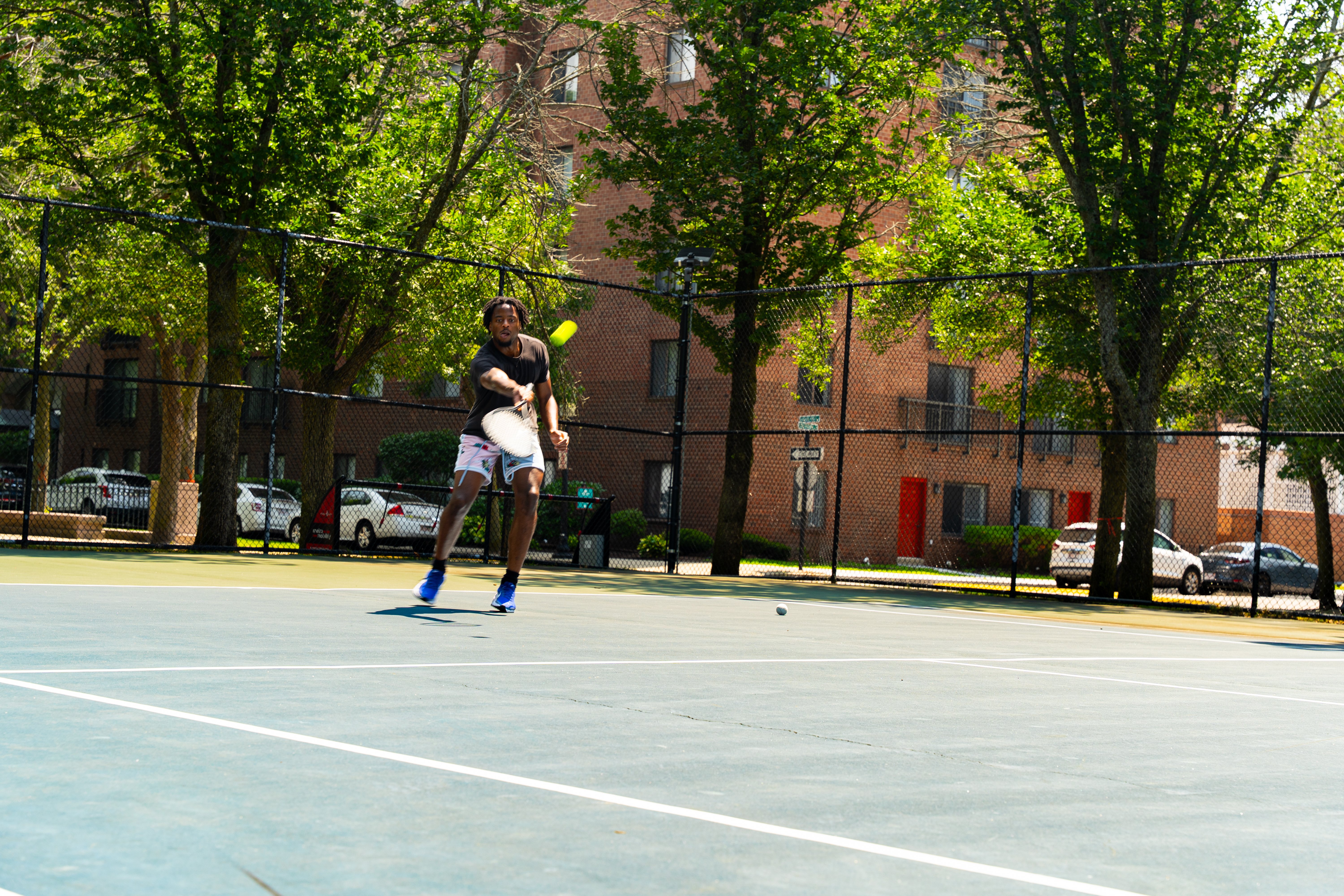 Tennis Player on Court A player is hitting a tennis ball on an outdoor court surrounded by trees and buildings.