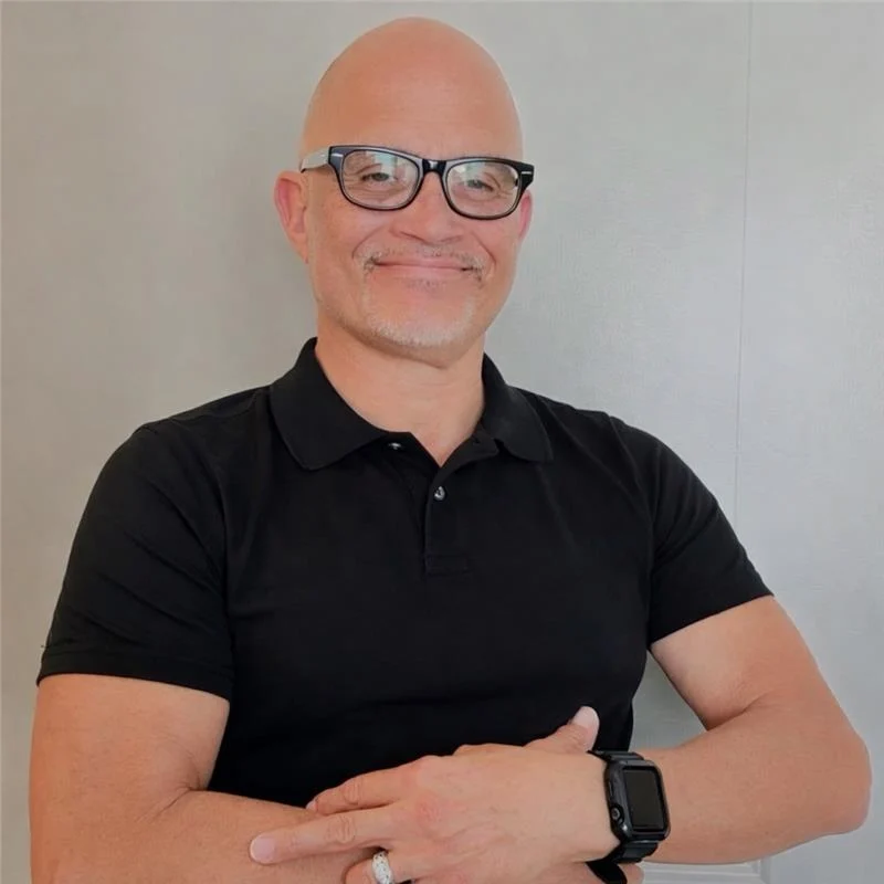 Confident Man Smiling A smiling man with glasses wearing a black shirt, arms crossed, sits against a plain background.