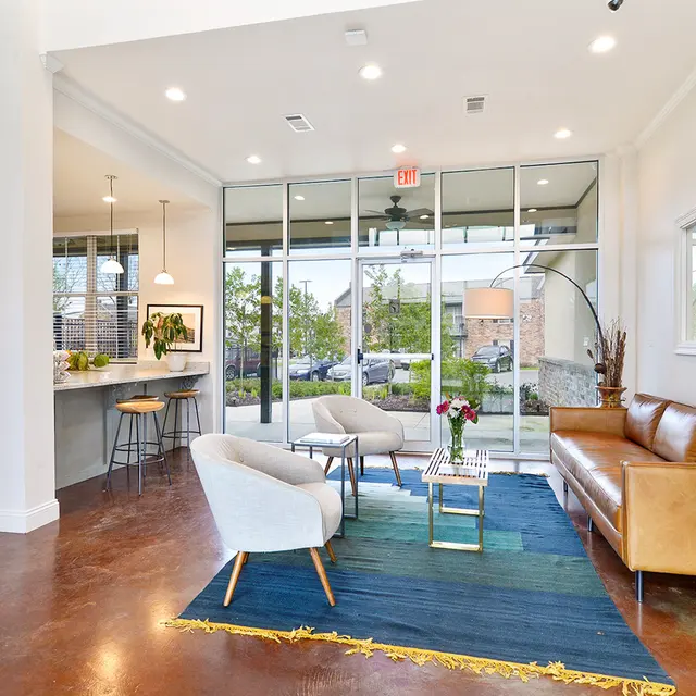 A bright and modern living space with a brown leather sofa, light-colored armchairs, and a coffee table on a blue and yellow area rug. Large windows allow natural light to fill the room, showcasing a view of greenery outside.