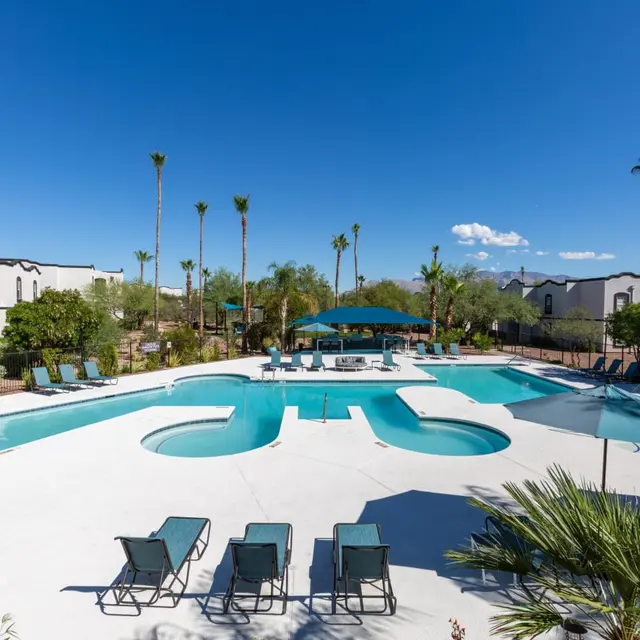 A spacious outdoor pool area featuring a large swimming pool, a jacuzzi, and several lounge chairs surrounded by palm trees and greenery under a clear blue sky.