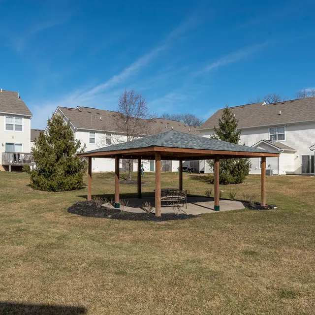 A spacious backyard featuring a wooden pavilion with a stone floor surrounded by green grass, small conifer trees, and residential buildings in the background under a clear blue sky.