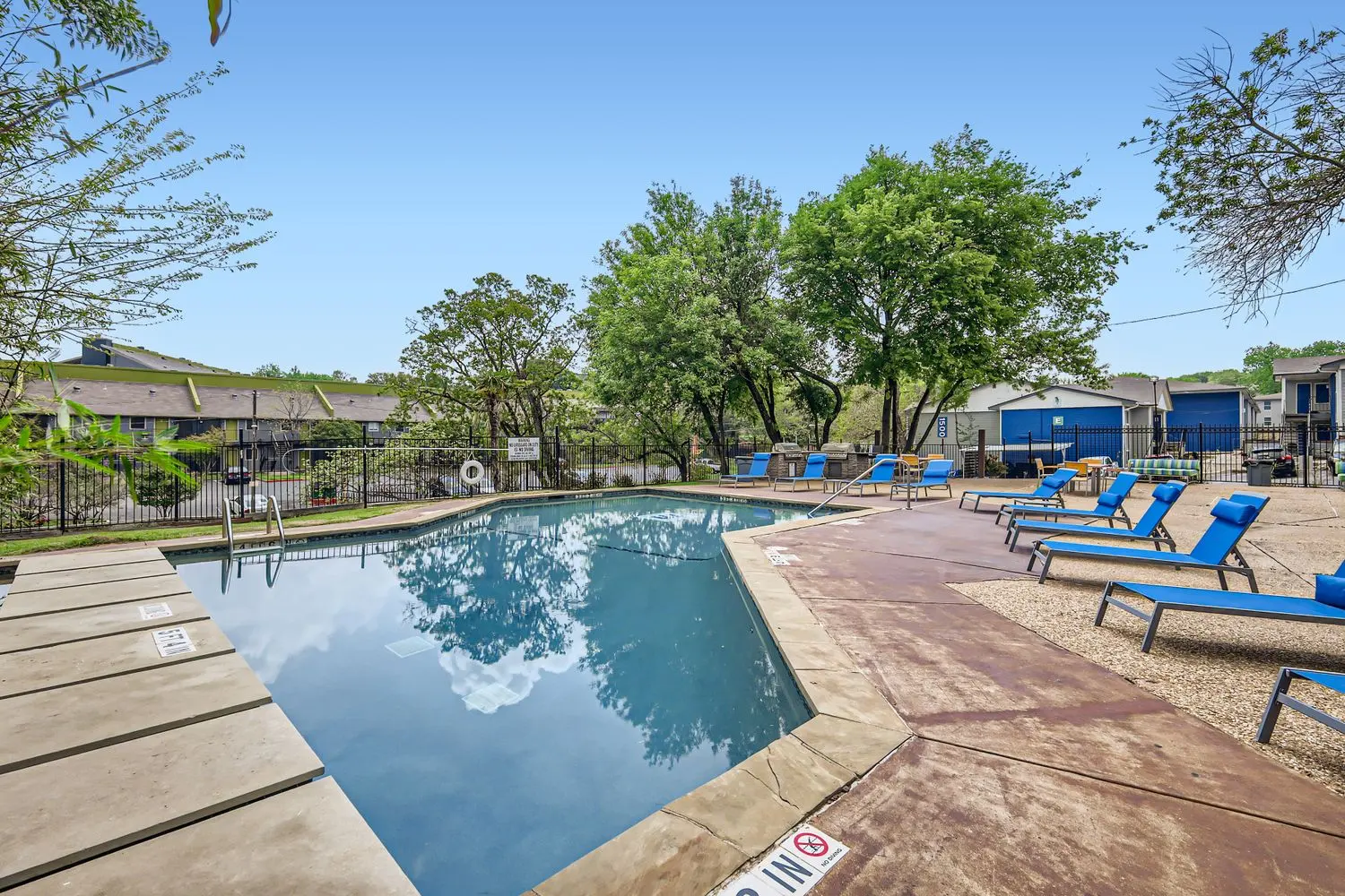 A residential pool area featuring a clear water swimming pool surrounded by blue lounge chairs under green trees.