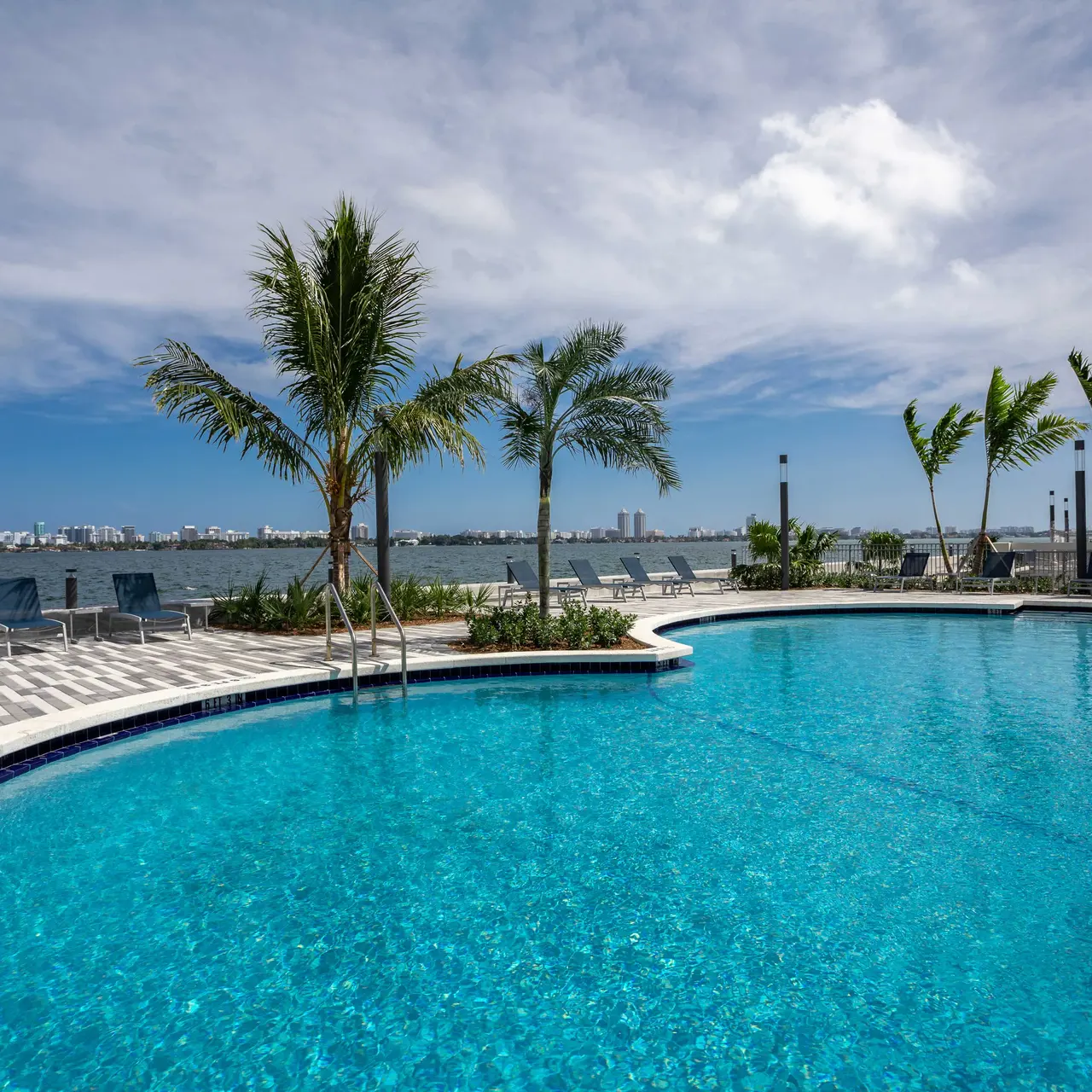 A luxury swimming pool with clear blue water surrounded by palm trees and lounge chairs, overlooking a body of water and a city skyline in the distance under a partly cloudy sky.