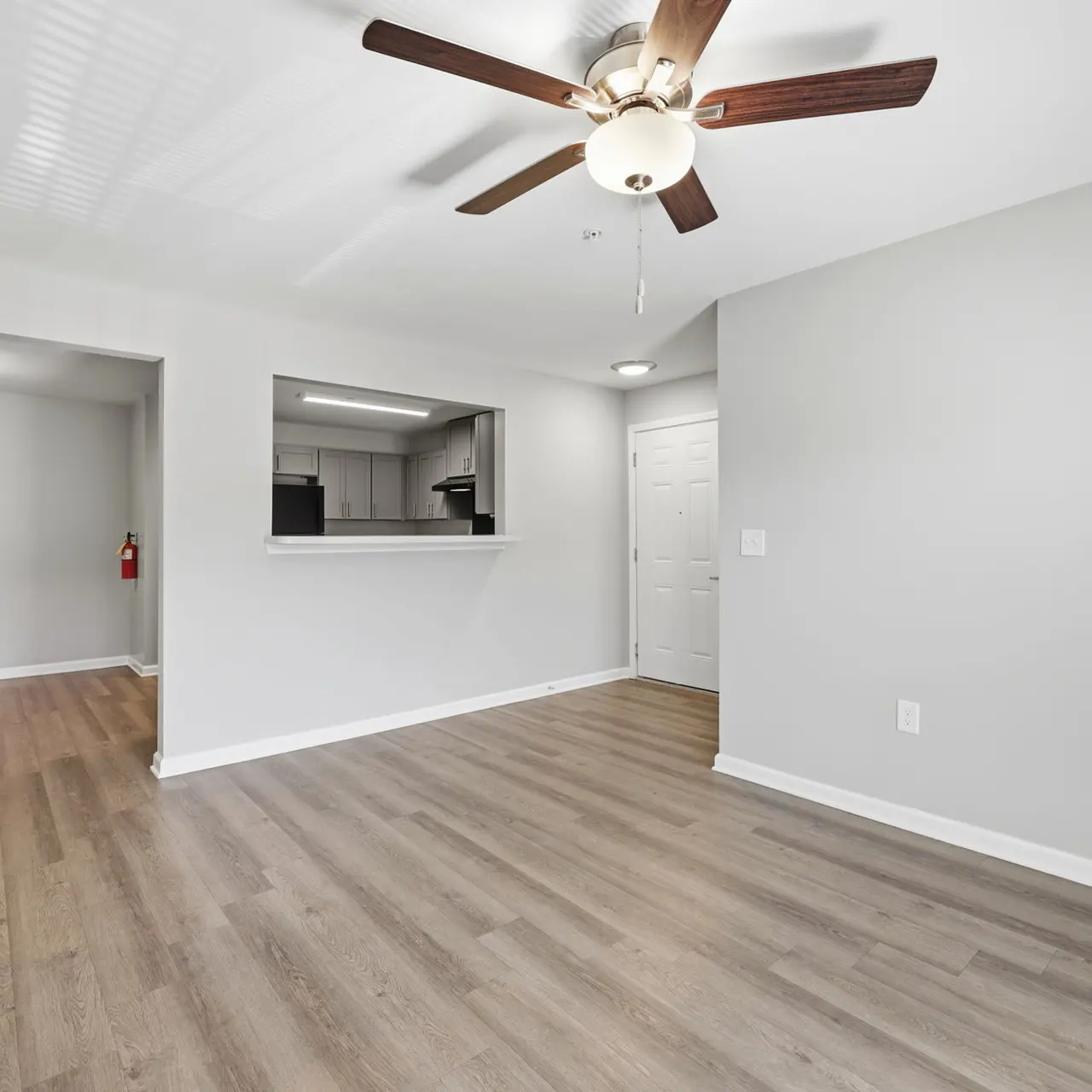 Interior view of a modern apartment featuring a living space with wooden flooring, a ceiling fan, and a view of a kitchen through an opening.