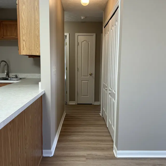 Interior view of a hallway leading from a kitchen, featuring light wooden flooring and beige walls. A door is visible at the end of the hallway, with an adjacent closet. Overhead lighting provides illumination.