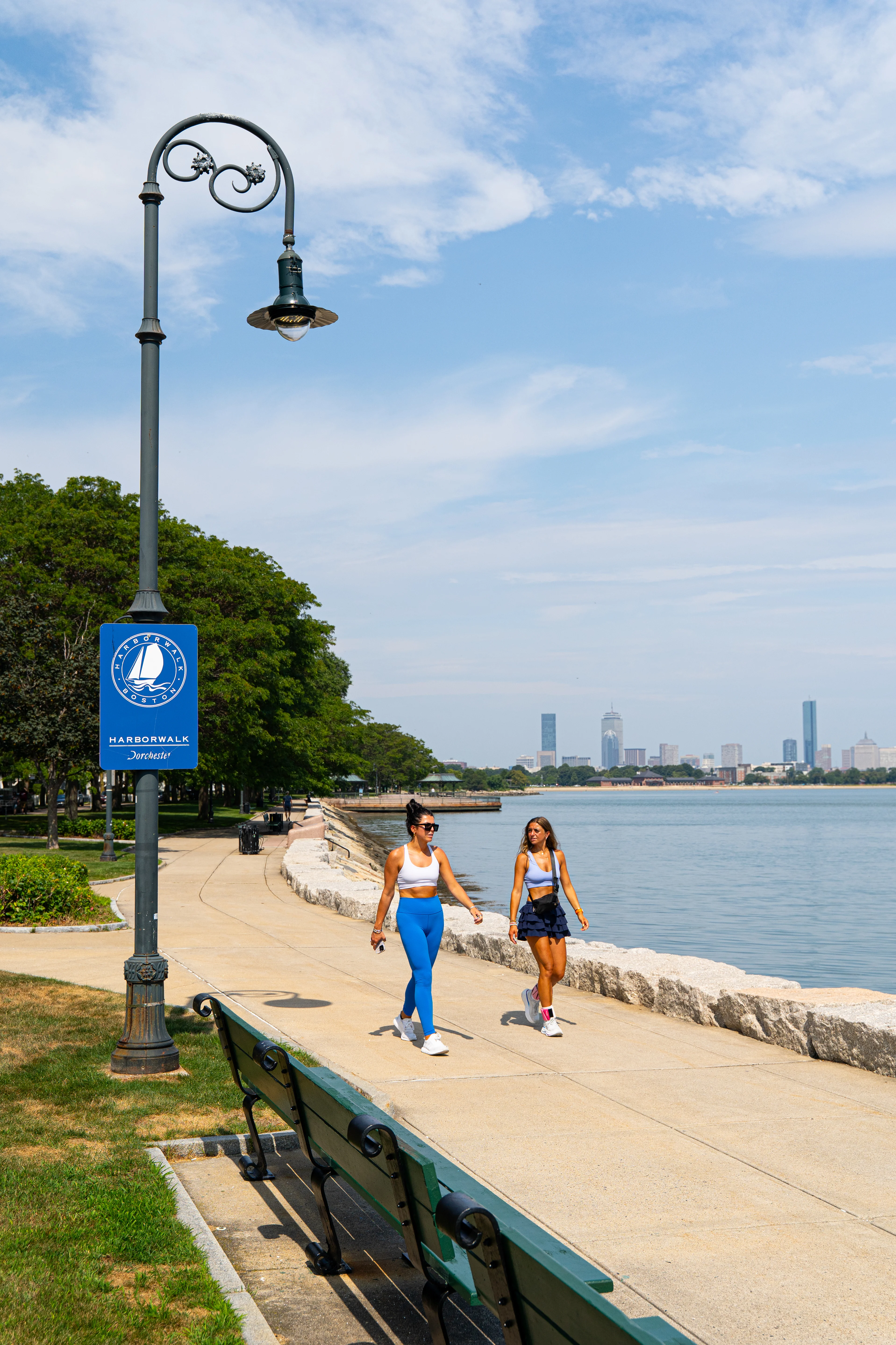 Lakeside Walking Path Two women walking along a lakeside path with benches and a lamp post in view, city skyline in the background.