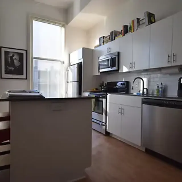 A modern kitchen with white cabinetry, stainless steel appliances, and large windows providing natural light. There's a small table with red chairs and some books on the kitchen counter.