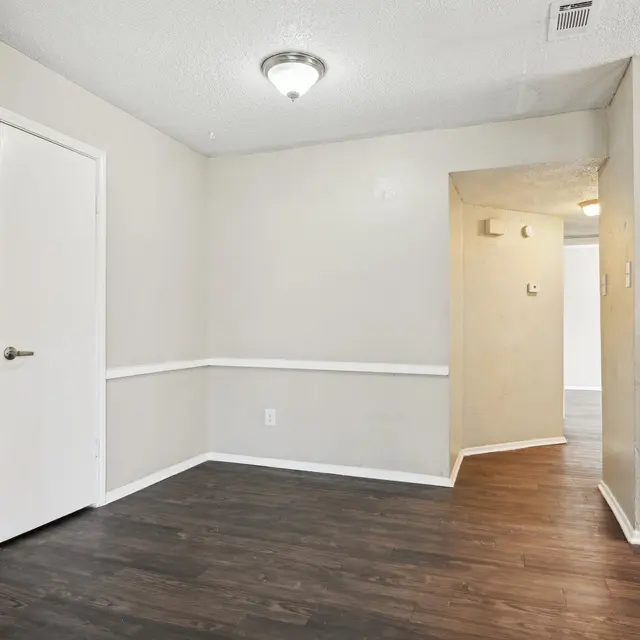 A small, empty corner of a room with light-colored walls and dark wooden flooring. There is a white door on the left and an entrance to another room on the right. The ceiling has a light fixture, creating a bright atmosphere.