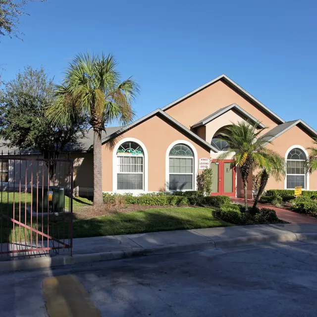 A single-story house with a beige exterior and green accents, surrounded by palm trees and a manicured lawn. A gated entrance is visible in the foreground.