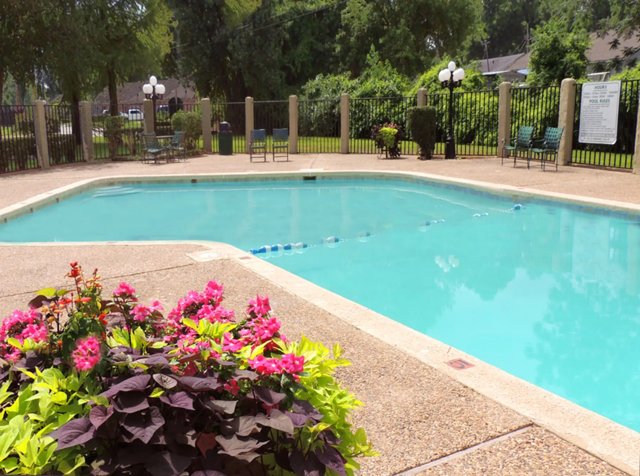 A clear swimming pool surrounded by vibrant flowers and a fenced area with chairs and lighting fixtures.