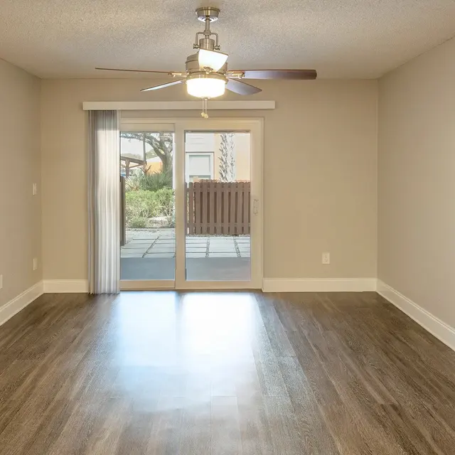 An empty living room with a ceiling fan, hardwood flooring, and a sliding glass door leading to an outdoor area.