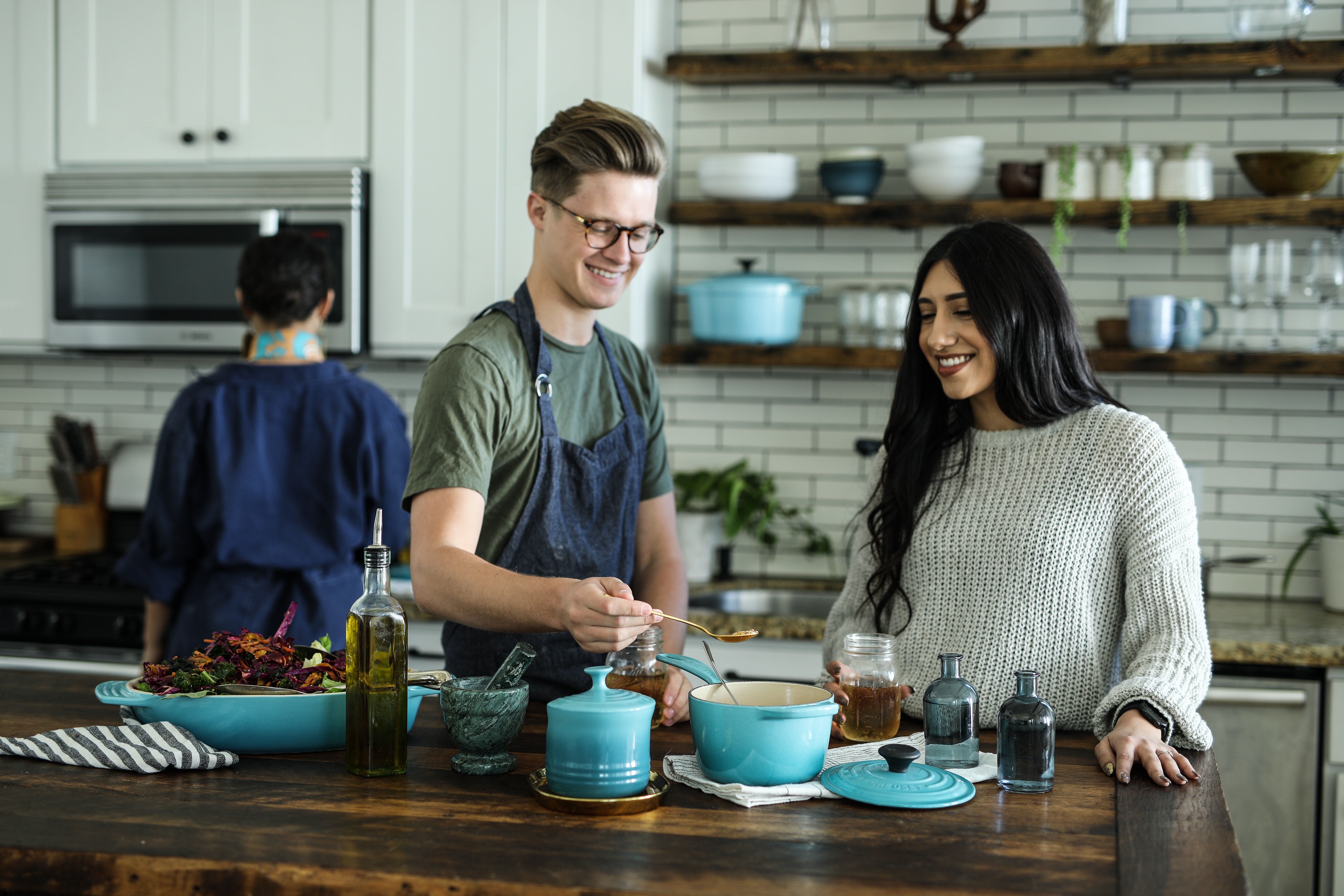 Main Street Apartments A joyful scene in a modern kitchen where a man and a woman are interacting while preparing food. The man, wearing an apron, is stirring a pot, and the woman is smiling and watching him. In the background, another person is working in the kitchen. The kitchen features wooden shelves and stylish kitchenware.