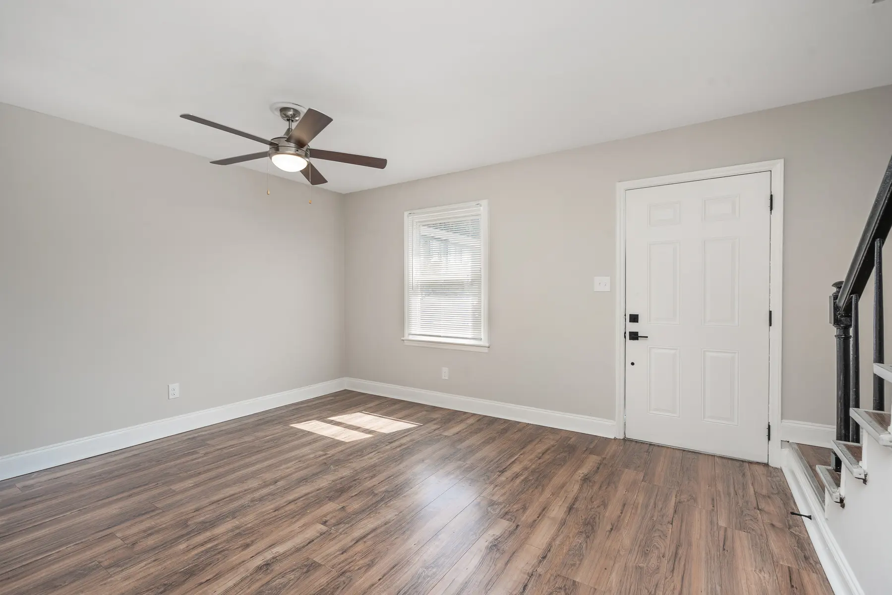 An empty living room with light-colored walls and a ceiling fan. There is a window allowing natural light and a door that leads outside.