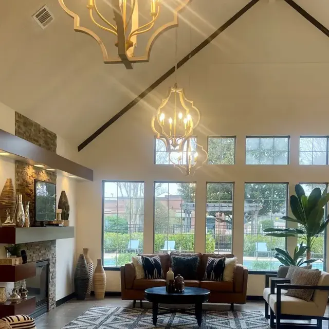 A spacious and modern living room featuring high ceilings, large windows, and stylish lighting fixtures. The room is furnished with a cozy sofa, patterned rug, and decorative elements on the walls and shelves.