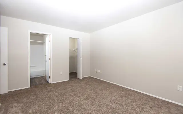 A spacious empty room with beige walls and carpeted flooring. There are two doors leading to closets, with one door partially opened revealing a closet space. The lighting is soft and there are no furnishings in the room.