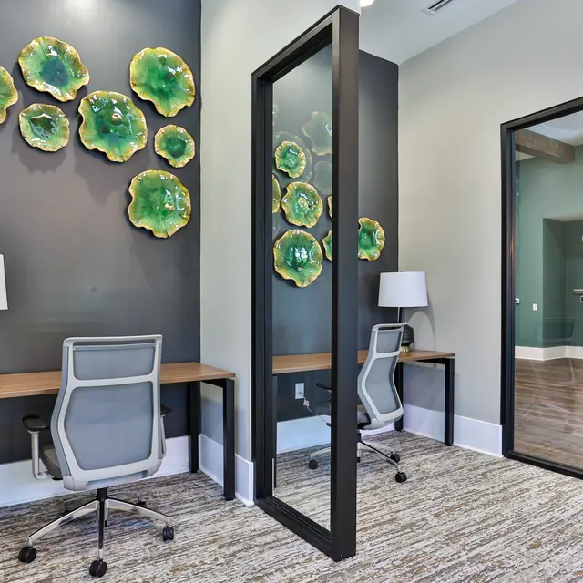 A modern home office featuring two workstations with ergonomic chairs and a decorative wall art composed of green circular shapes. The office has a dark accent wall and a large glass panel separating the space.