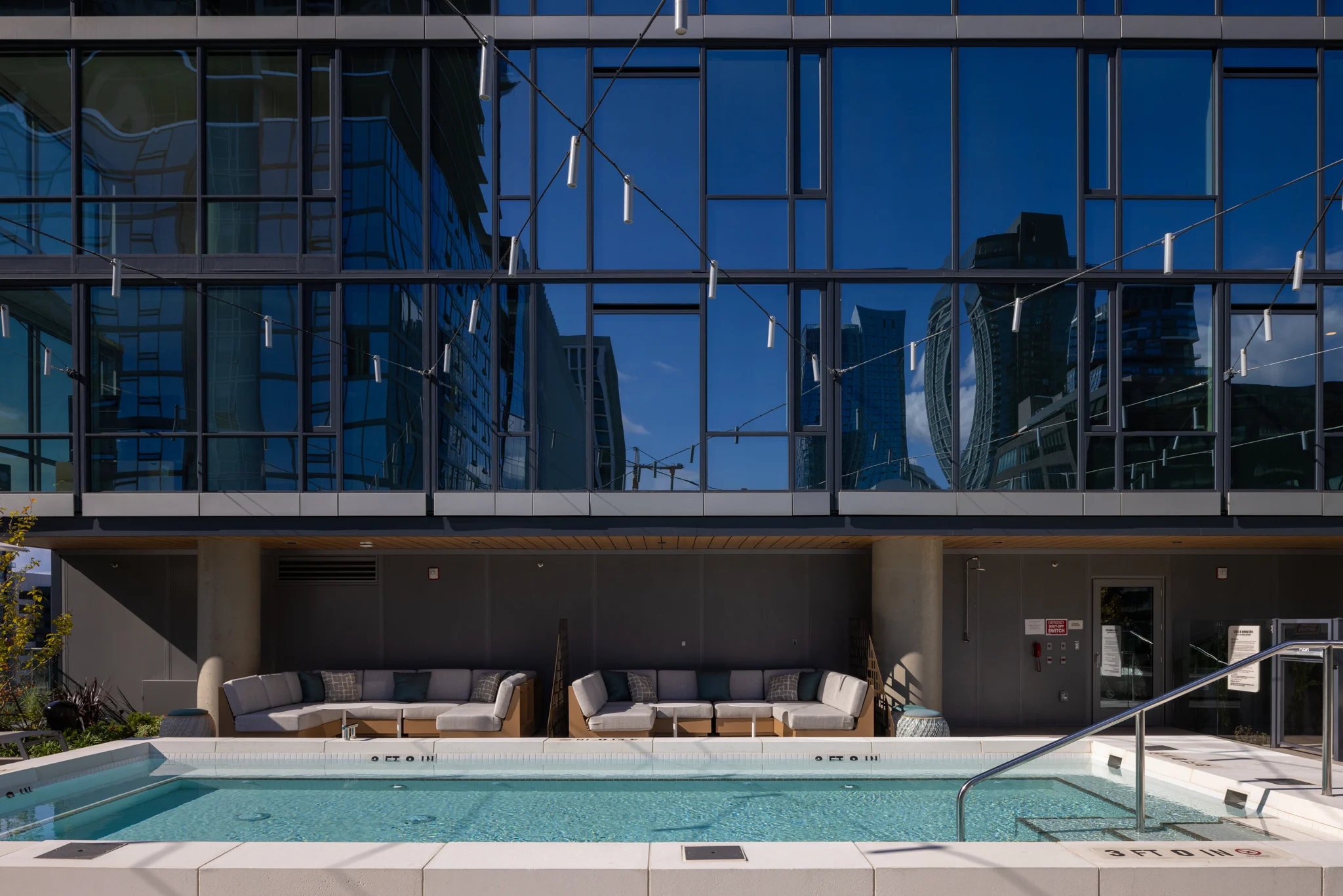 A sleek outdoor pool with a clean, modern design situated in front of a large glass building. The water is clear, and there are comfortable loungers nearby. The building's reflections can be seen in the glass facade.