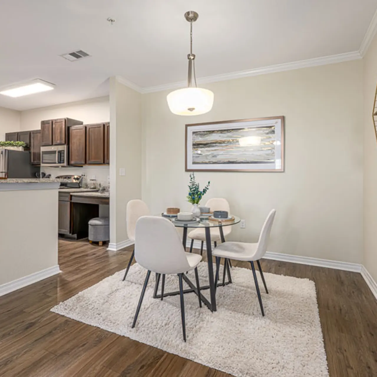 A modern dining area featuring a round table with four chairs, a vase of flowers, and a light fixture above. The kitchen is visible in the background with dark wood cabinetry and appliances. Light-colored walls and a plush rug enhance the inviting atmosphere.