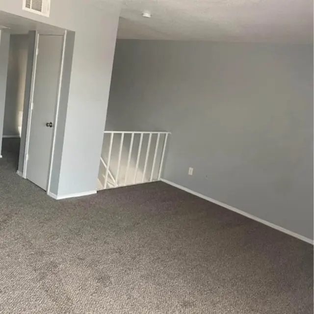 An empty room featuring gray walls and a carpeted floor. A ceiling light is visible, and there are doors on the left leading to another area. A staircase railing can be seen in the corner.