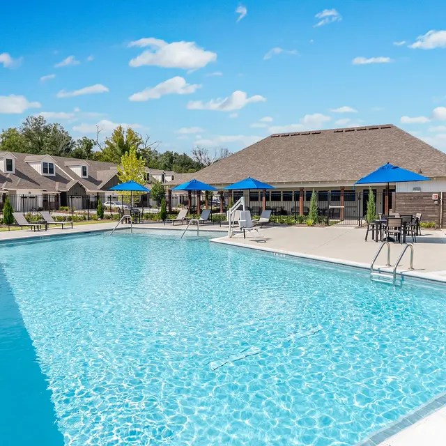 Outdoor Swimming Pool at Apartment Complex A large outdoor swimming pool surrounded by lounge chairs and umbrellas, with trees and a building in the background.