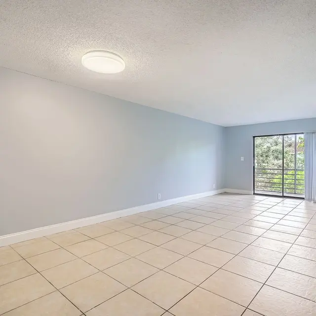 An empty room with beige tiled flooring, light gray walls, and a large window overlooking greenery.
