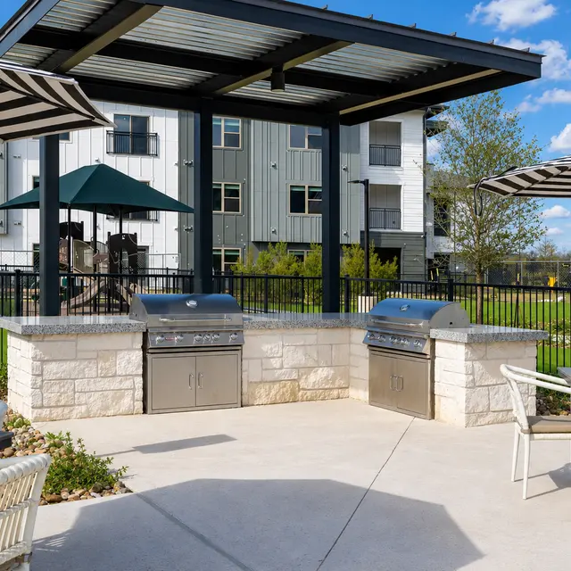 An outdoor grilling area with two stainless steel grills, surrounded by tables and chairs under striped umbrellas. In the background, there are residential buildings and a playground area.