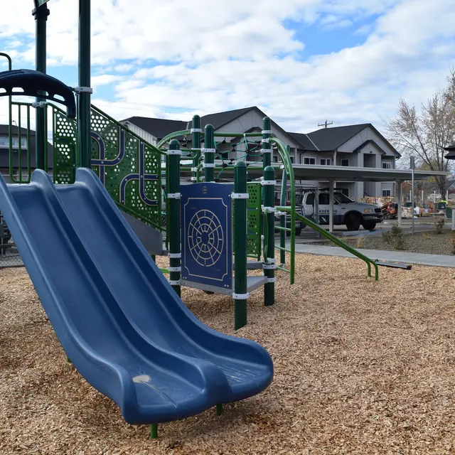 Playground Area with Slide A blue playground slide situated in a play area filled with wood chips. The slide is connected to a green climbing structure in the background, and residential buildings are visible in the distance under a cloudy sky.