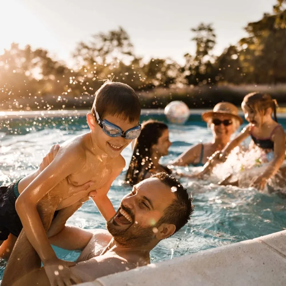 A joyful scene in a swimming pool with a man playing with a boy wearing goggles. The man is laughing, and the boy is sitting on his shoulders. In the background, women and children are enjoying splashing water and playing. The setting is sunny with trees around, capturing a warm, cheerful summer atmosphere.