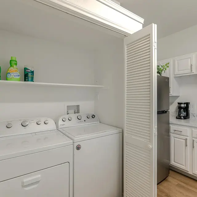A laundry room featuring a washer and dryer set, with laundry detergent and fabric softener on a shelf above. A kitchen area is visible in the background with white cabinets, a coffee maker, and a stove.
