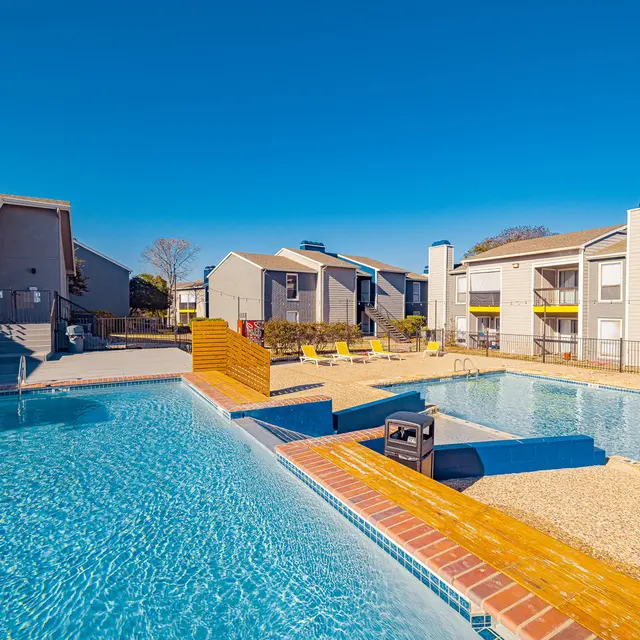 View of a swimming pool area with multiple pools, wooden decking, and surrounding apartment buildings under a clear blue sky.