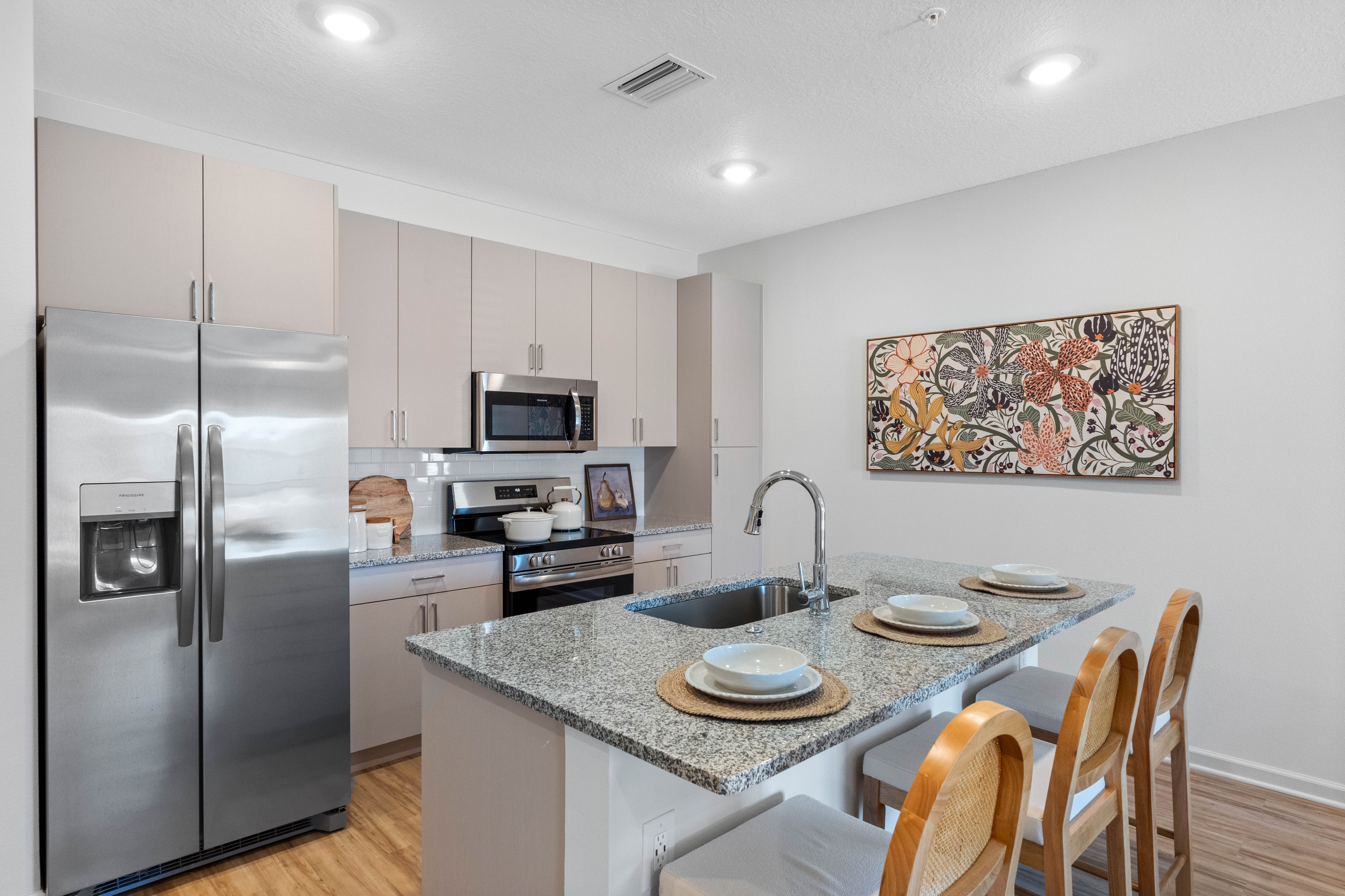 Modern Kitchen Interior A modern kitchen featuring sleek stainless steel appliances, light colored cabinets, and a granite countertop. A decorative wall art is displayed above the counter with three place settings visible.