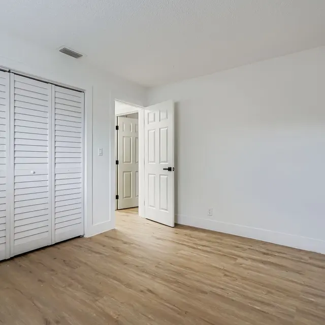 An empty room with wooden flooring, white walls, and a closet with shutter doors on the left side. A door leading to another room is visible on the right side.