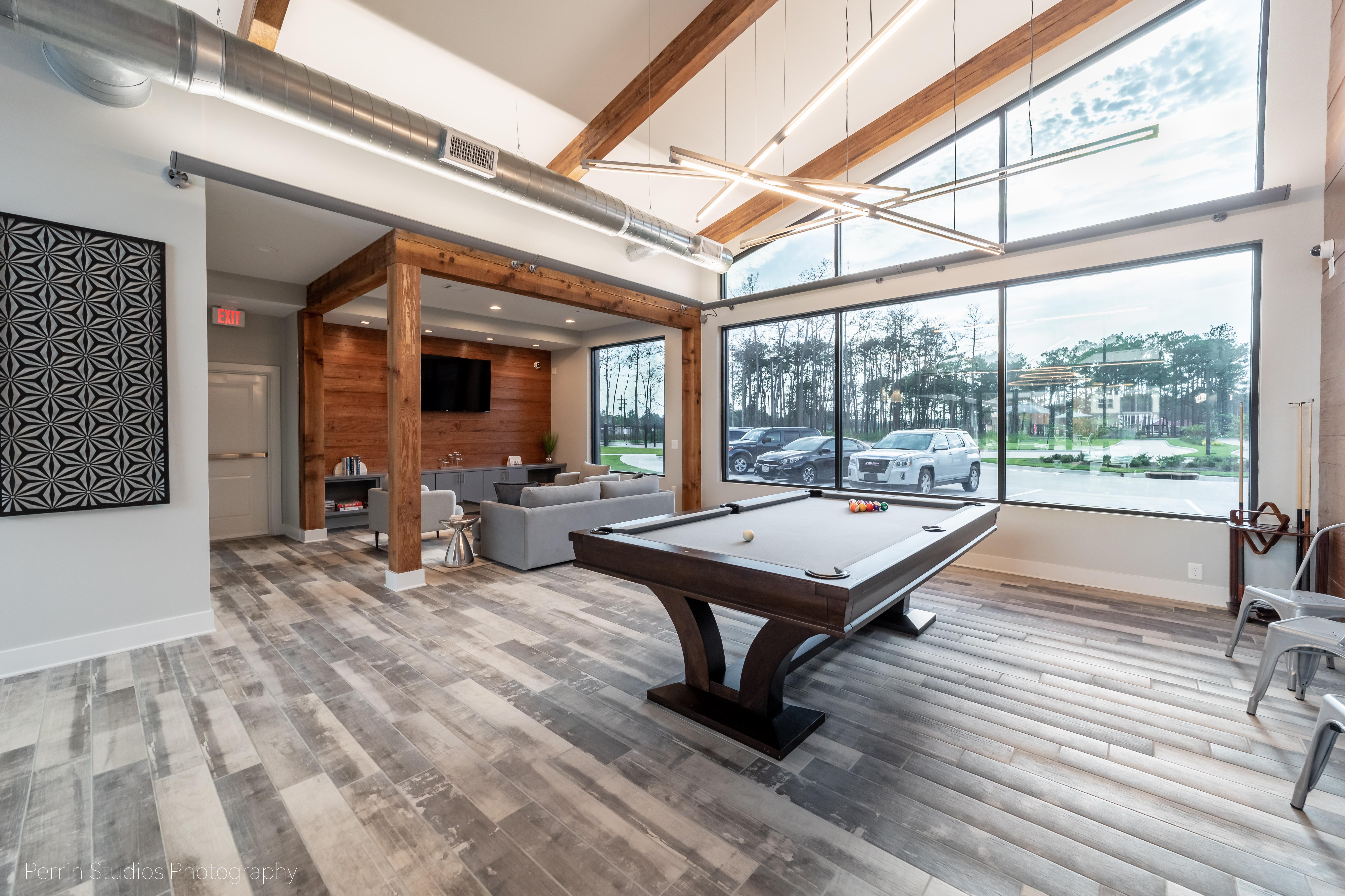 A modern game room featuring a pool table in the center, large windows letting in natural light, and a cozy seating area with gray couches and a TV mounted on the wall.