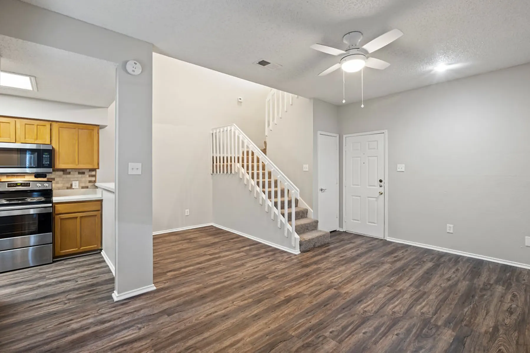 A modern living room with hardwood-like flooring, a staircase leading to an upper level, and a kitchen area visible on the left with wooden cabinets and stainless steel appliances.