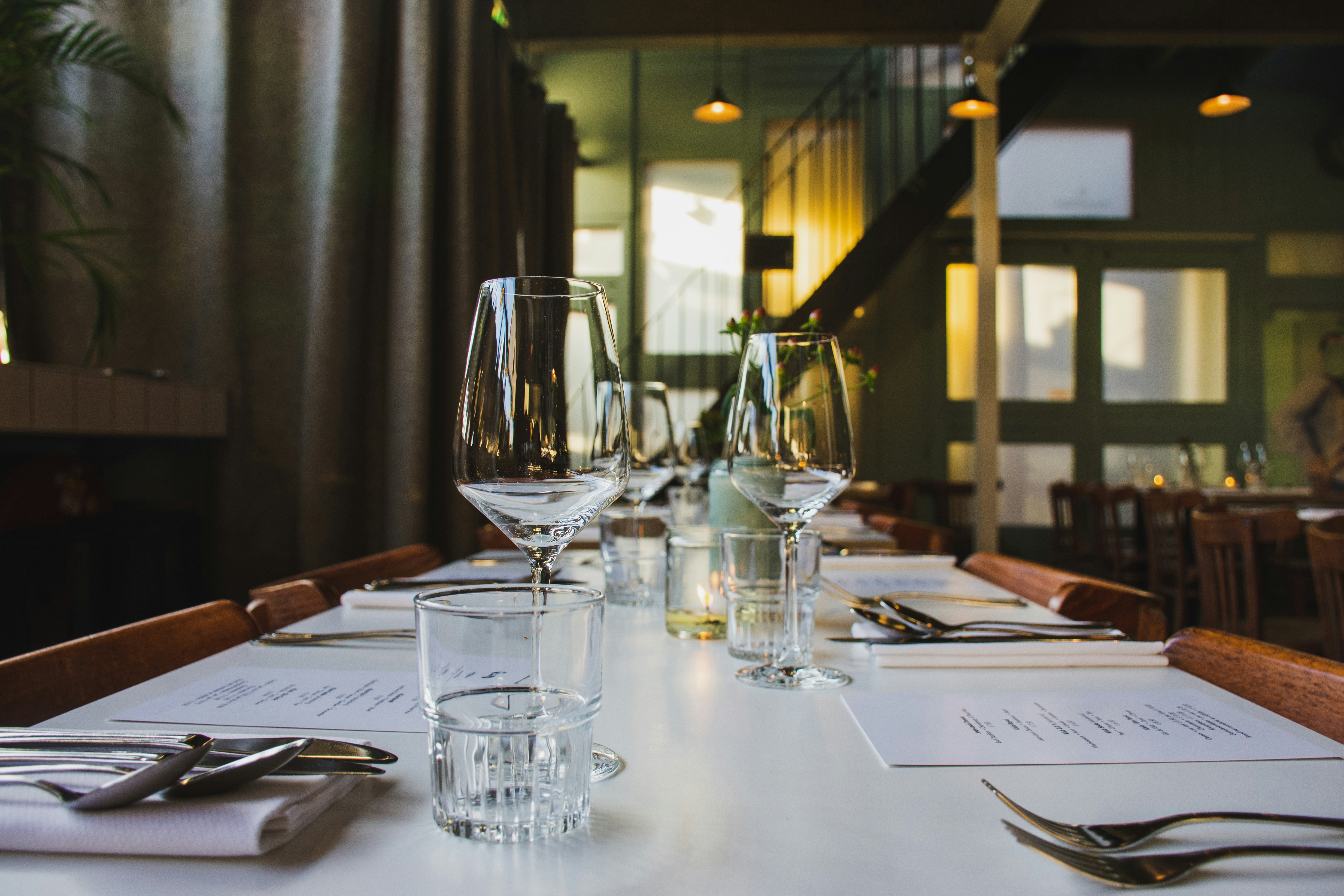 A beautifully arranged dining table in a restaurant, featuring glassware, cutlery, and menus on a white tablecloth. The ambient lighting creates a warm atmosphere, with wooden chairs and a staircase visible in the background.