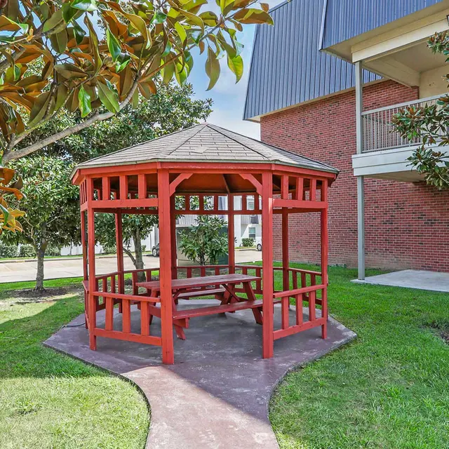 A red gazebo with a tiled roof nestled on a lawn surrounded by greenery, with a gray building in the background.