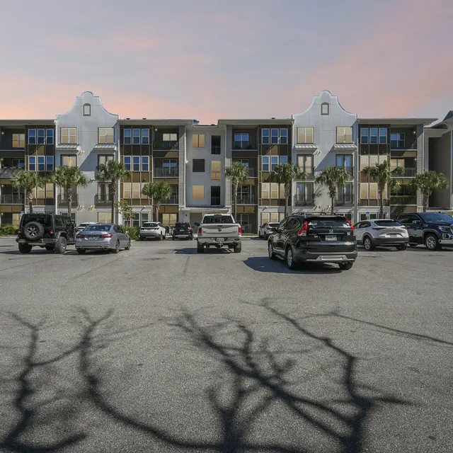 A view of a modern apartment complex with a parking lot in the foreground. The building features balconies and palm trees around it, under a sky with a gradient of colors.