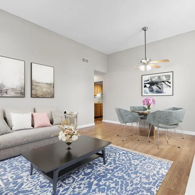 A modern living room featuring a light gray wall, a comfortable sofa with pink and white cushions, a dark coffee table, and a blue patterned rug. In the background, there's a dining area with a round table and chairs, along with a glimpse of a kitchen.