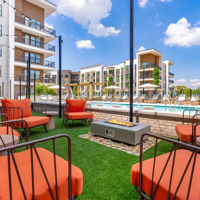 A lounge area with bright orange chairs facing a swimming pool surrounded by modern apartment buildings under a cloudy blue sky.