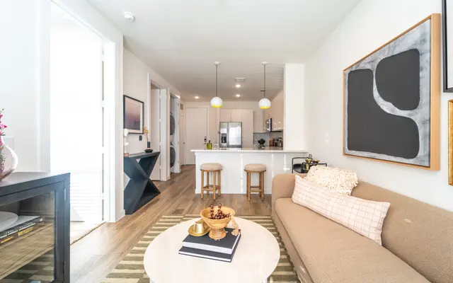 Stylish Modern Living Room A modern living room with a light-colored sofa, a round coffee table, and a small kitchen in the background. The room features artwork on the walls and tasteful decor on the coffee table and furniture.